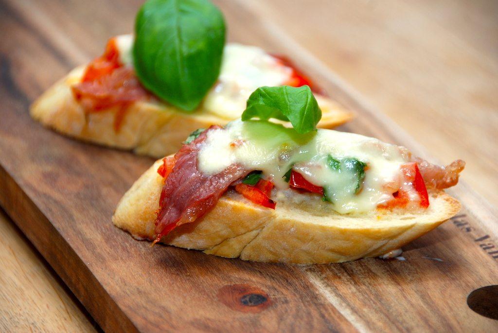 Bruschetta med mozzarella er en skøn forret fra Italien, der dog sagtens også kan nydes uden ost. Smør brødet med hvidløgsolie, og læg så de øvrige ingredienser oven på, inden bruschettaen bages i ovnen. Foto: Guffeliguf.dk.