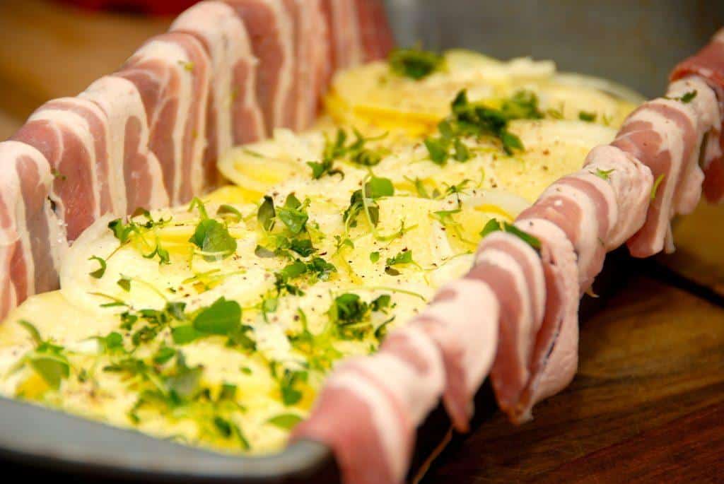 En form smøres grundigt med olie eller smør, hvorefter baconskiverne lægges med et lille overlap, og enderne lægges op over kanten. Fyld derefter lagvis med kartofler, løg, hvidløg og timian og der pensles med piskefløde. Foto: Madensverden.dk.